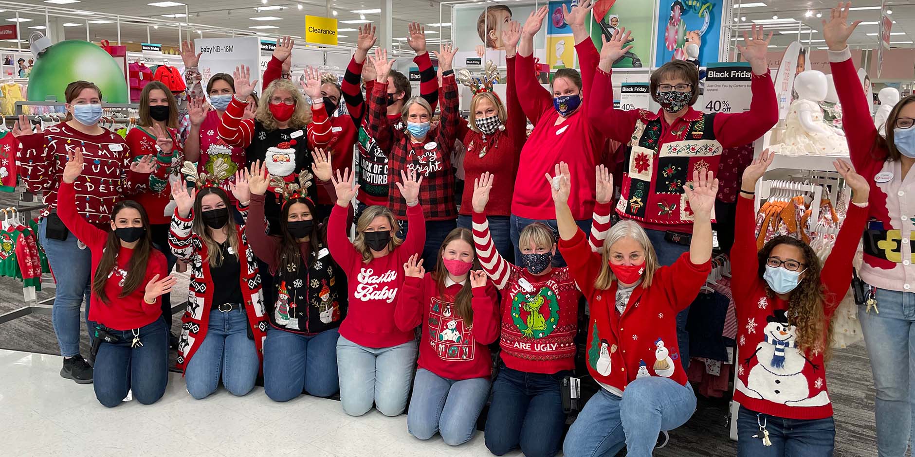 "A large group of team members stand or kneel together with their hands in the air in front of a Target holiday display. They're all wearing festive sweaters and face masks."
