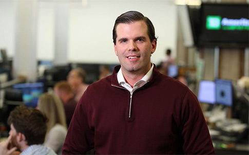 A team leader posing in front of his colleagues in an open office, smiling and wearing a smart business casual outfit