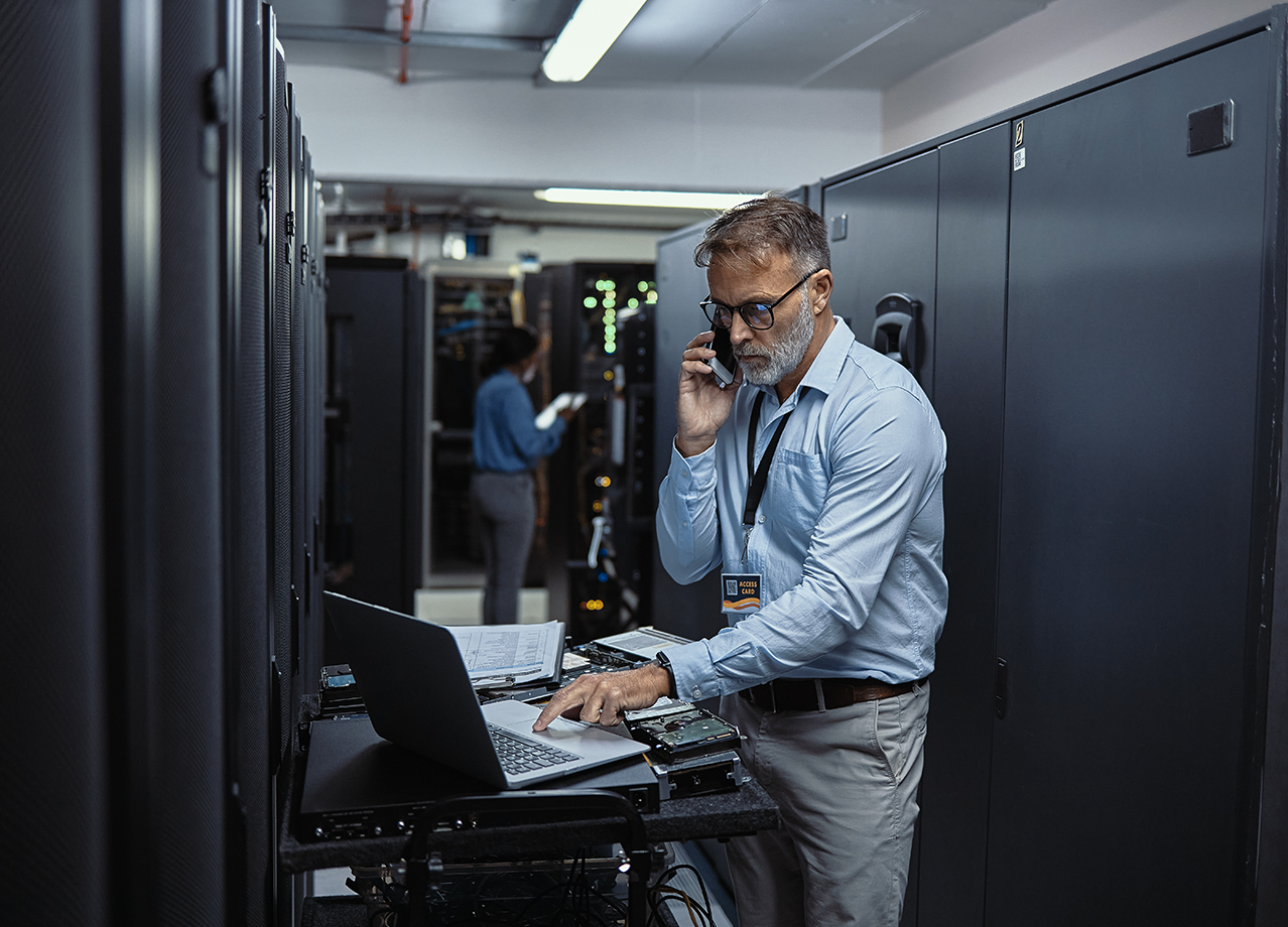 Employee working on a laptop and speaking on a phone in a data center.
