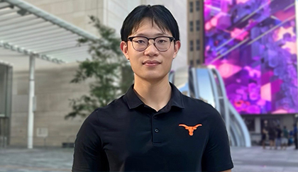Person in a black collared shirt and glasses standing in an urban plaza with buildings, trees, and colorful digital displays in the background.
