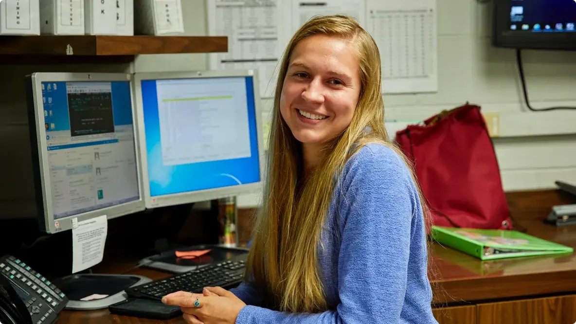 A woman smiling while sitting at a desk in front of two computer monitors.