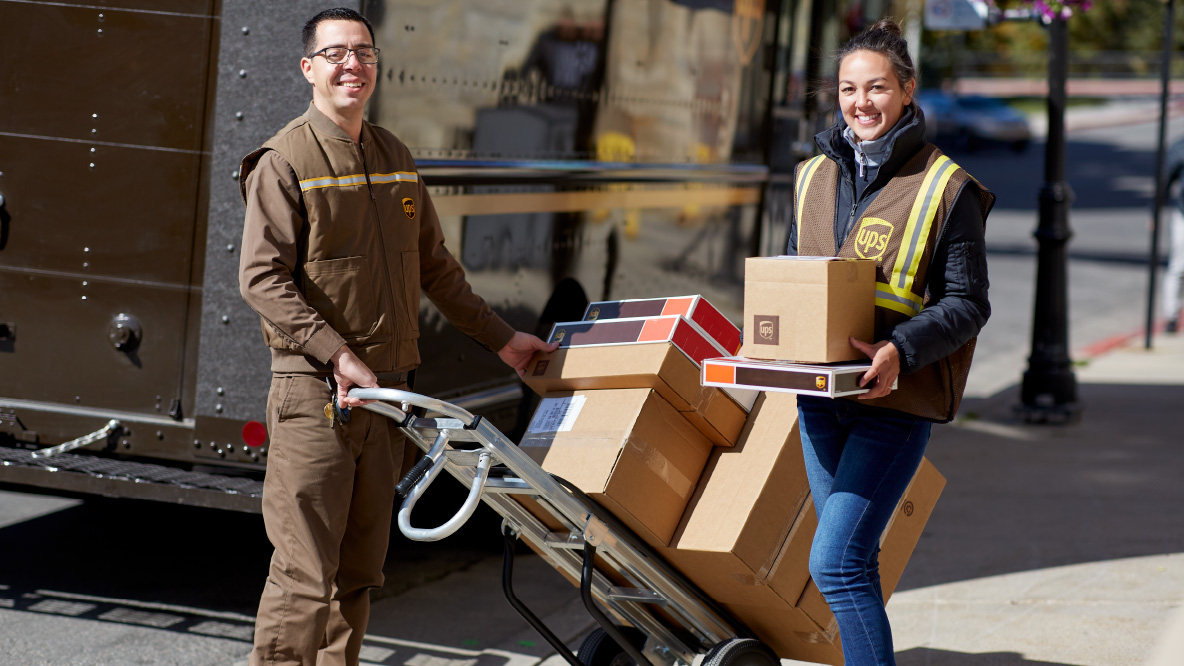 Two UPS workers, a male with a dolly filled with packages and a female holding two packages