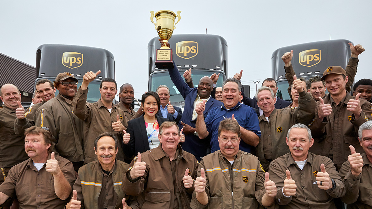 A group of smiling UPS workers with their thumbs up, in front of 3 UPS semi trucks, holding up an award