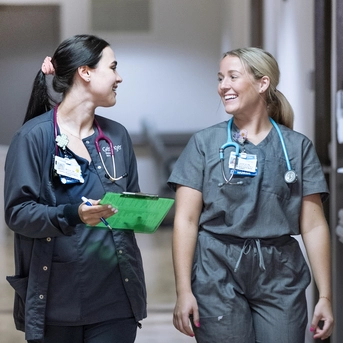 Two healthcare professionals in scrubs walking through a hallway while reviewing information on a clipboard.