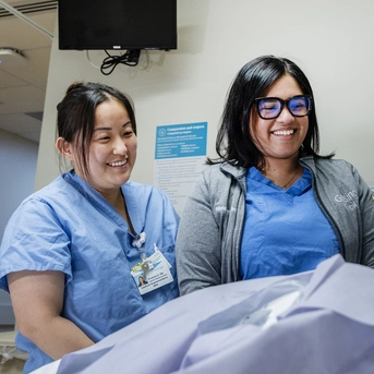 Two healthcare team members in scrubs review information together beside a patient bed in a clinical care setting.