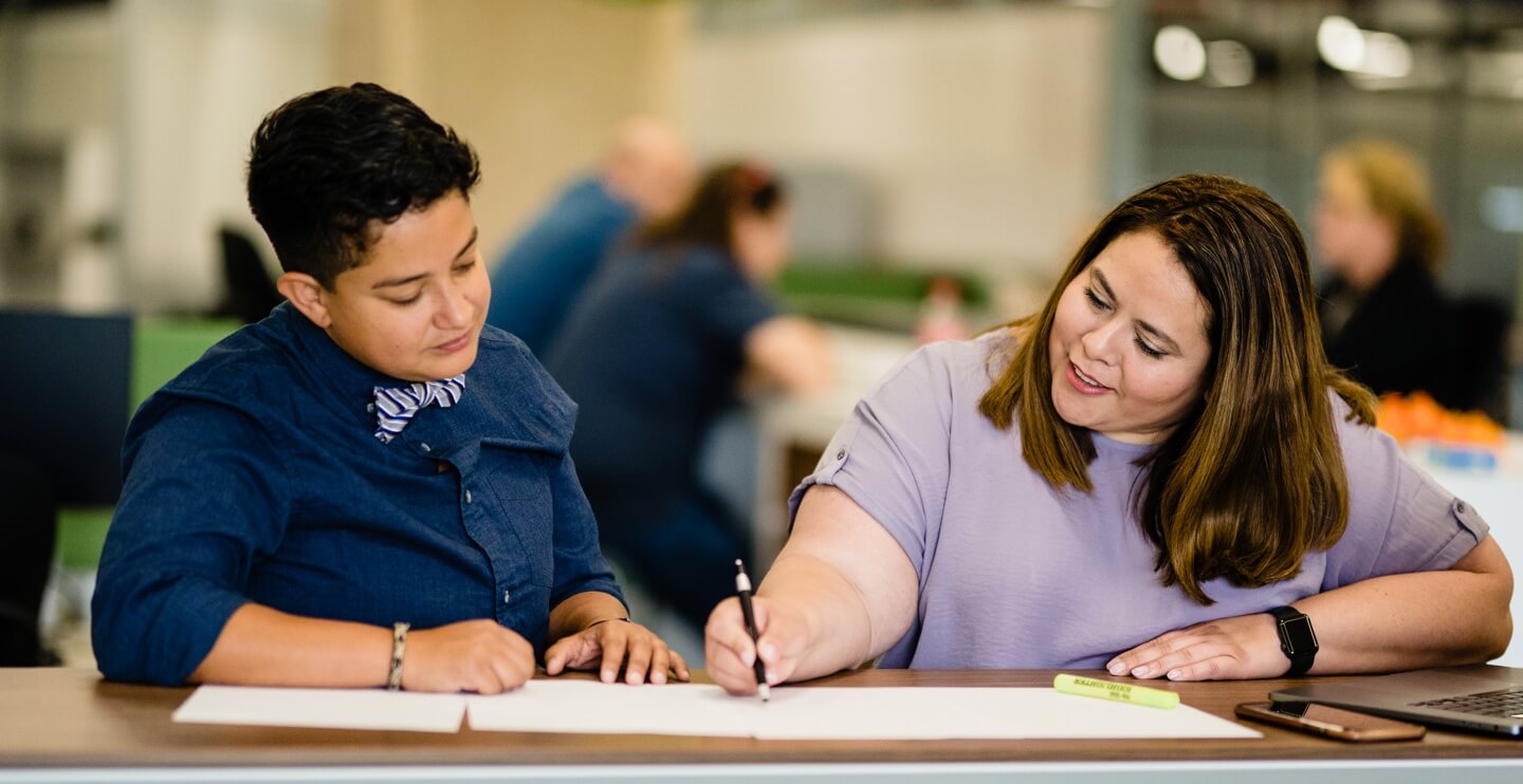 A woman sitting next to another person and pointing to a piece of paper with a pen