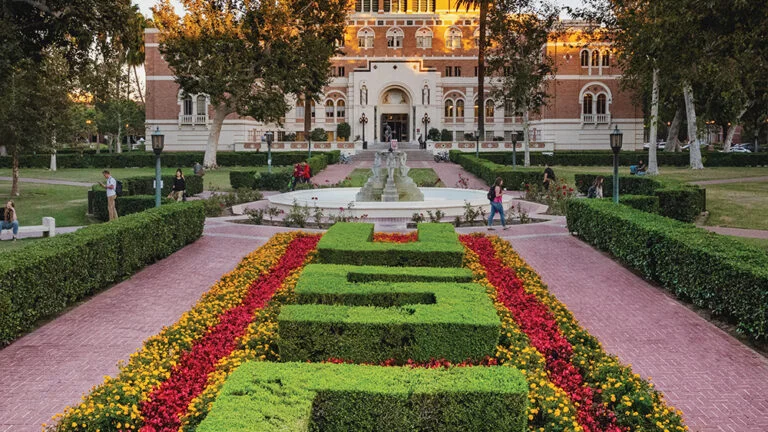 Landscaped garden at a university campus with hedges trimmed to spell “USC,” colorful flower beds, a central fountain, and a large historic brick building in the background.