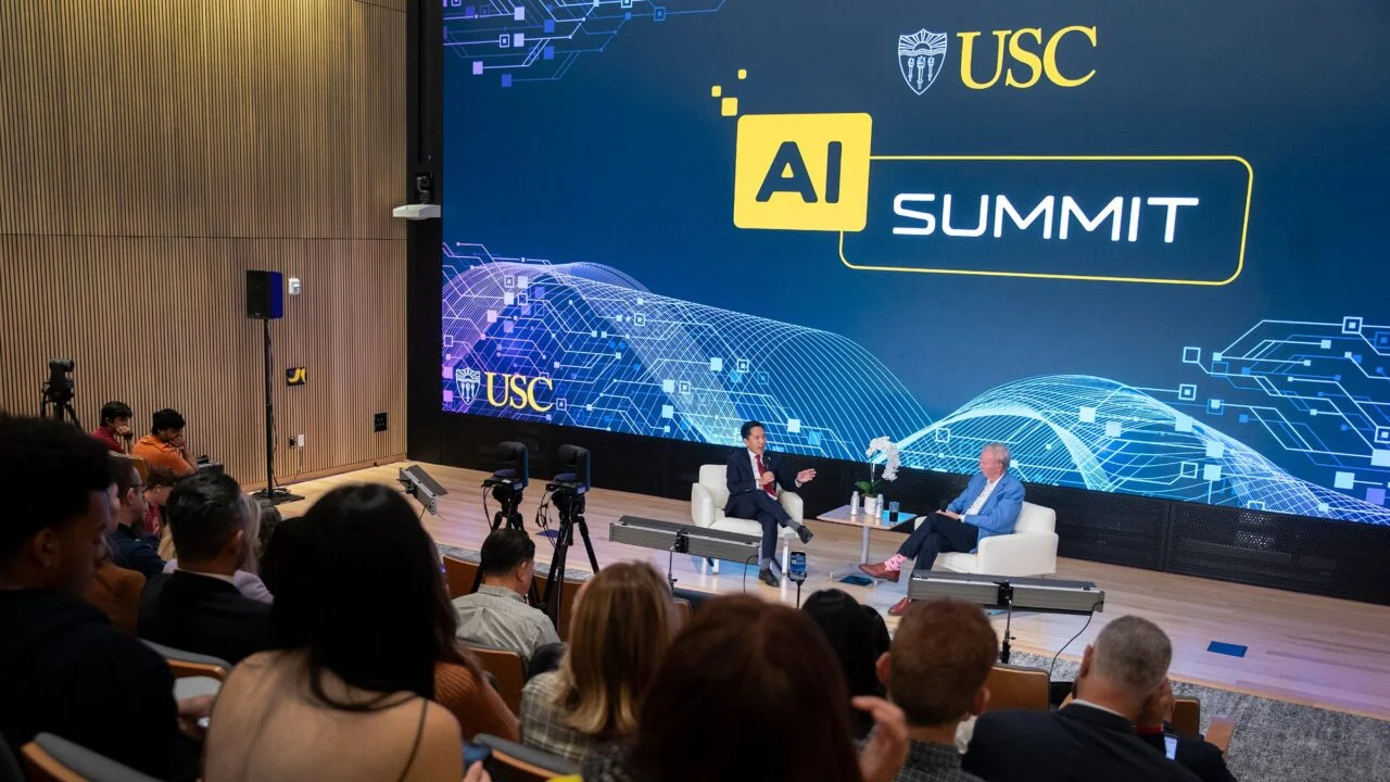 Two speakers seated on stage at a USC AI Summit event, speaking in front of a large screen displaying “AI Summit” and the USC logo, with an audience in the foreground.