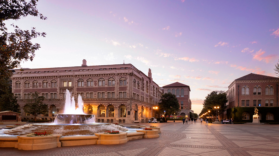 Wide view of a university campus plaza at dusk, featuring a large illuminated fountain in the foreground and historic brick-and-stone academic buildings lining a broad pedestrian walkway. Warm lights glow from the arches and streetlamps, while a pastel pi