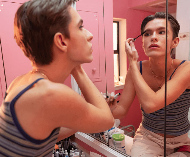 Young gender non-conforming person applies makeup in their home bathroom mirror