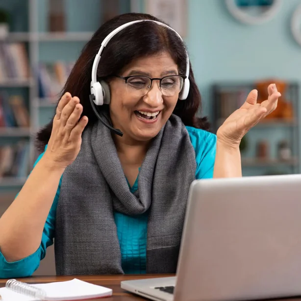 A middle-aged woman wearing headphones is gesturing excitedly during a video call in front of her laptop in a home office setting.