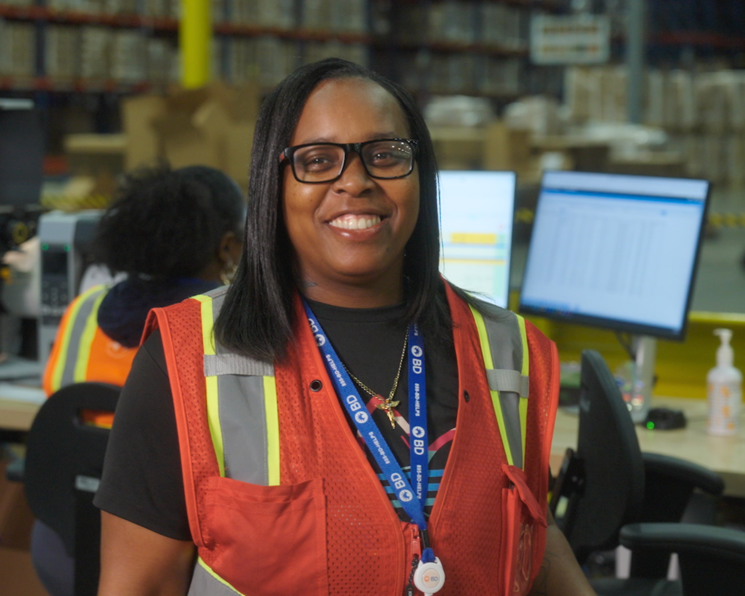 Empleado de BD con chaleco de seguridad en un puesto de trabajo dentro de un almacén, con otra persona trabajando en una computadora al fondo.