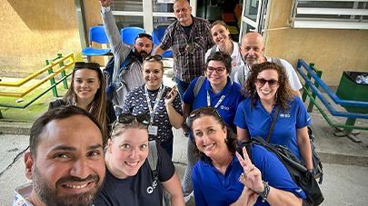 A group of individuals standing closely together outside a building entrance, many holding up peace signs or making group gestures. The background includes outdoor seating, railings, and a doorway.