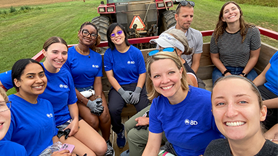 A group of individuals seated in the back of an open tractor trailer during an outdoor activity, all wearing BD-branded shirts or casual clothing. Fields and farm equipment are visible in the background.