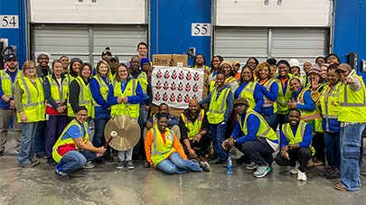A large group of individuals wearing safety vests gathered inside an industrial facility, standing in front of loading dock doors labeled with numbers. The group poses together around a display board featuring a pattern of red and white icons.