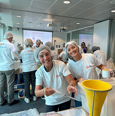 Individuals wearing BD Volunteer shirts, hairnets, and gloves working together in an indoor room with tables of packing materials and supplies, with two individuals in the foreground preparing items and smiling toward the camera.