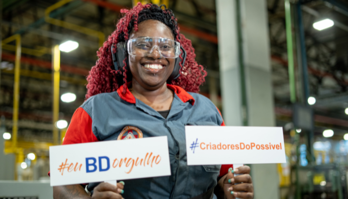 Black woman in manufacturing plant holding up signs which read Proud to be BD