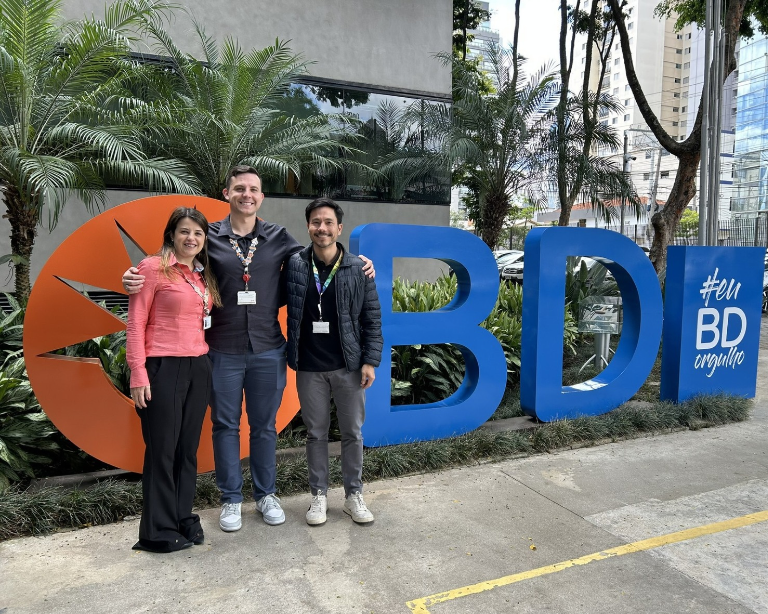 Three associates posing in front of a large BD sign on a landscaped campus.