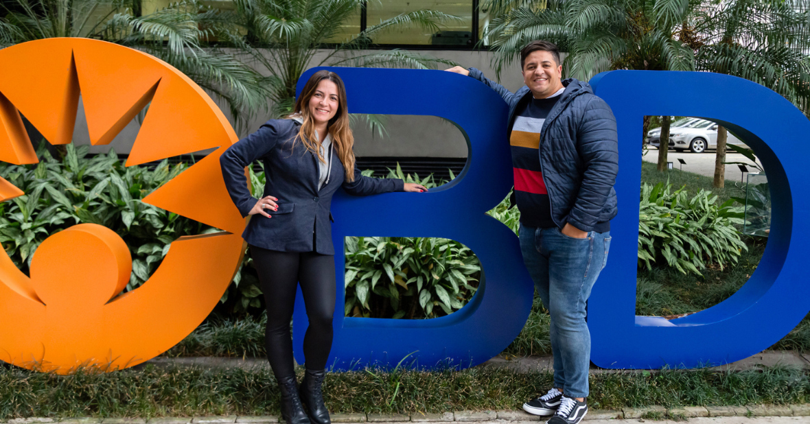 Dois associados da BD posam ao ar livre em frente ao logotipo da BD, em um ambiente corporativo com áreas verdes.