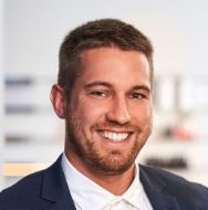An individual smiling with short brown hair and a trimmed beard poses in a navy suit and white shirt, with a softly blurred office background.