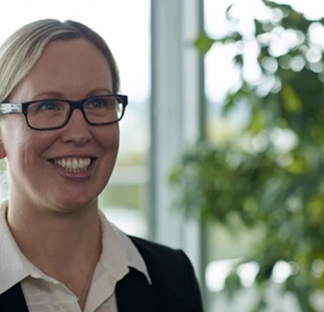 Una persona con gafas y vestimenta de negocios —que incluye una camisa de cuello de color claro y una chaqueta oscura— se encuentra de pie en un interior, con un fondo desenfocado que muestra ventanas y follaje verde.