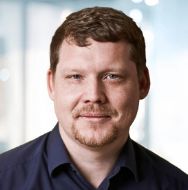 Professional headshot of an individual with short brown hair and a trimmed goatee in a navy shirt, looking calmly at the camera in an office background.