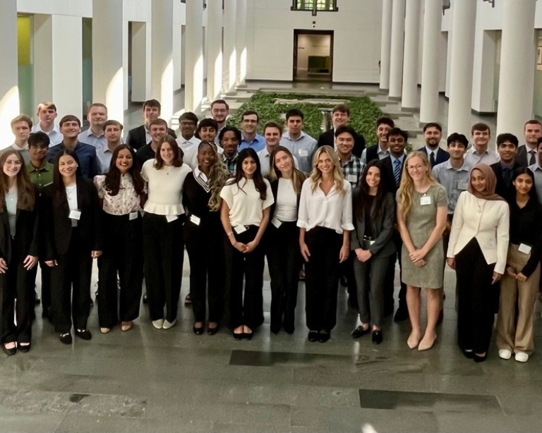 Un gran grupo de personas vestidas de negocios posando juntas en un espacioso atrio con altas columnas y luz natural.