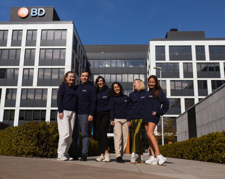 Grupo de personas que visten sudaderas azul marino a juego de BD, de pie frente a un moderno edificio de oficinas de BD de varias plantas, con grandes ventanales y arbustos ajardinados.