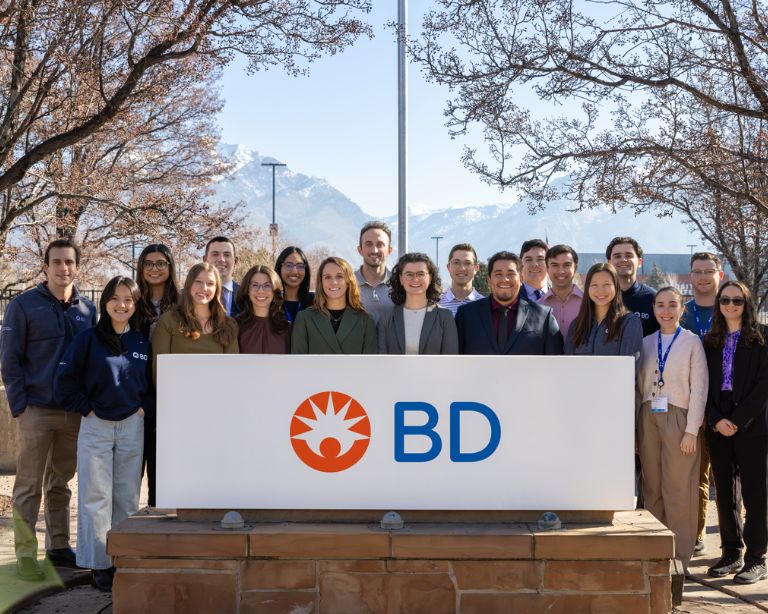 Group of individuals wearing matching navy BD sweatshirts standing in front of a modern multi‑story BD office building with large windows and landscaped bushes.