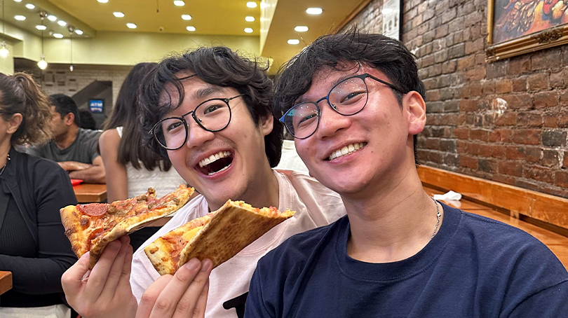Dos personas sentadas en un restaurante informal, sosteniendo grandes rebanadas de pizza, con paredes de ladrillo visto y asientos tipo cabina de fondo.