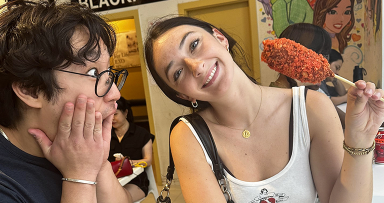 Dos personas de pie dentro de un local de comida informal; una de ellas sostiene un aperitivo crujiente en un palito.