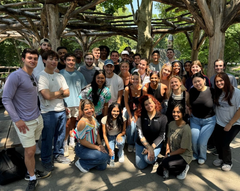 Un gran grupo de personas se reunió al aire libre, bajo una rústica pérgola de madera, rodeado de árboles y luz solar.