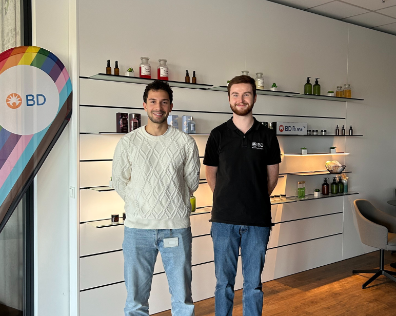 Dos personas de pie frente a una pared de exhibición de BD, con estantes que contienen diversas botellas y envases de productos, junto a un gran letrero con la marca BD y un diseño de arcoíris.