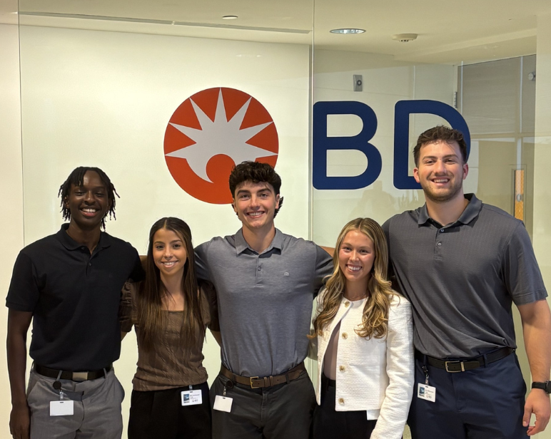 Grupo de personas de pie frente a un gran logotipo de BD, exhibido en una pared de cristal dentro de un entorno de oficina.