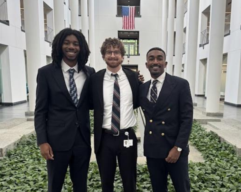 Tres individuos vestidos con trajes formales de pie en un gran atrio con altas columnas blancas y vegetación.