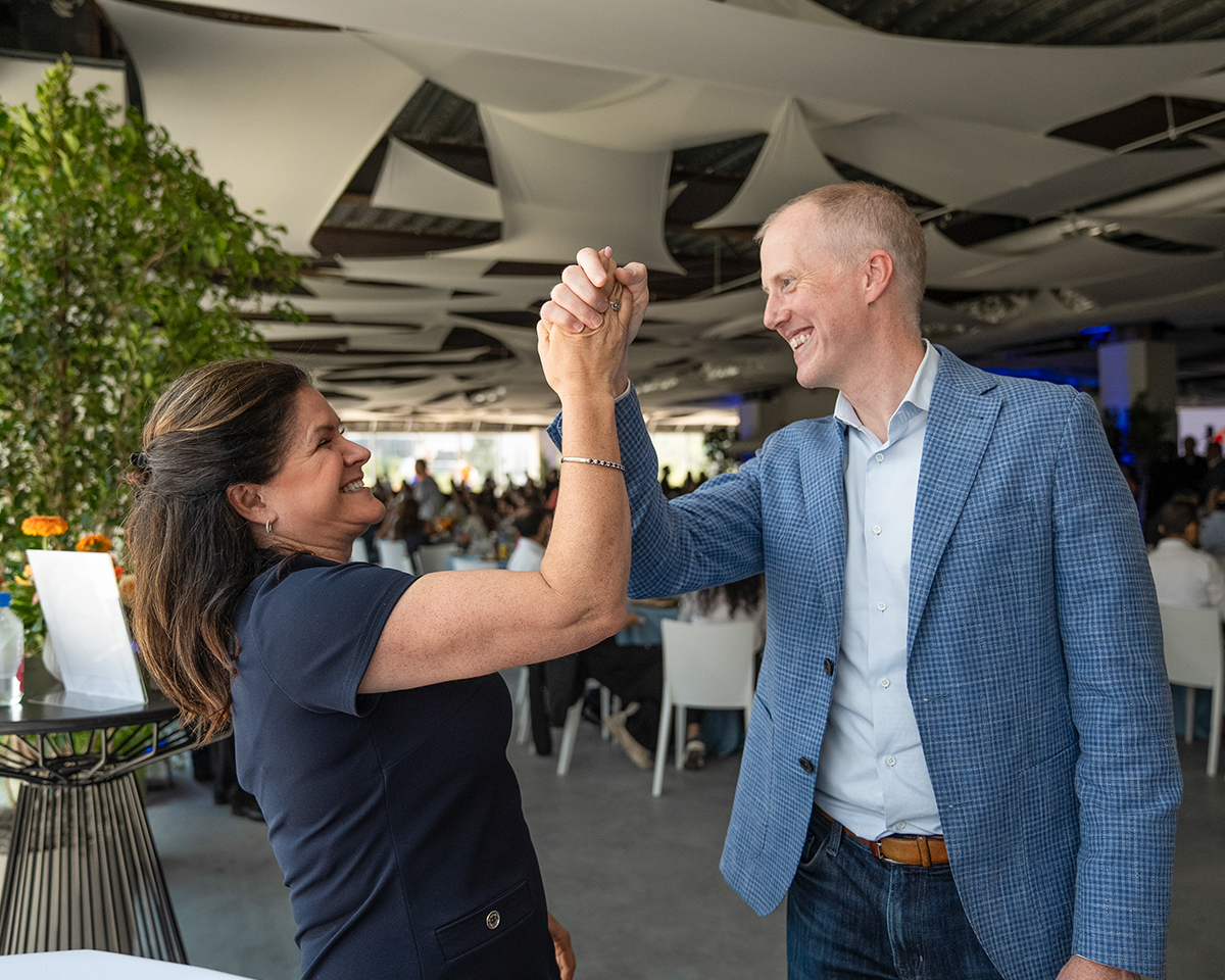 Dos individuos de pie en un espacio para eventos, alzando y uniendo las manos en un gesto de celebración o reconocimiento.