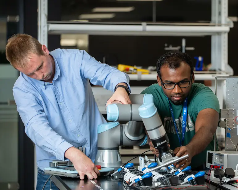 Dos individuos trabajando juntos para ajustar componentes en un brazo rob&oacute;tico dentro de un espacio de trabajo t&eacute;cnico.