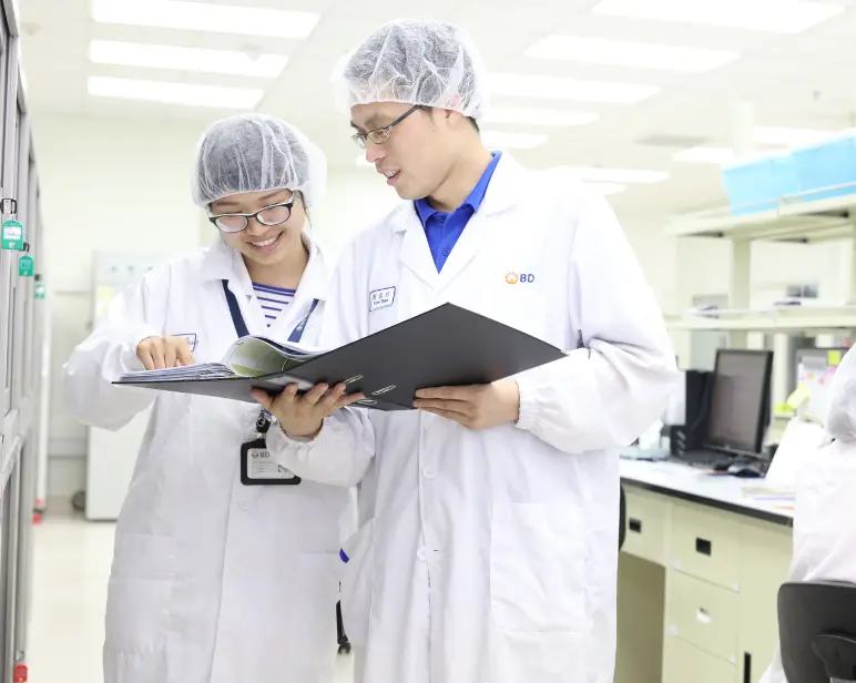 Dos personas, con batas de laboratorio de BD y cubrecabezas, revisando documentos juntas en el interior de un laboratorio limpio y luminoso.