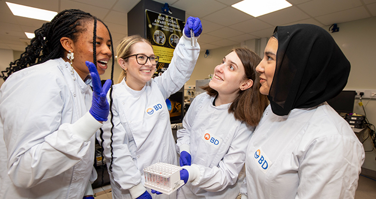 Cuatro individuos, con batas de laboratorio BD y guantes azules, examinando juntos un tubo de ensayo en el interior de un laboratorio de investigación brillantemente iluminado.