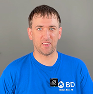 Persona con una camiseta azul de BD Bruker Bio Inc. y un peque&ntilde;o dispositivo electr&oacute;nico sujeto a la camiseta, de pie frente a un fondo gris liso.