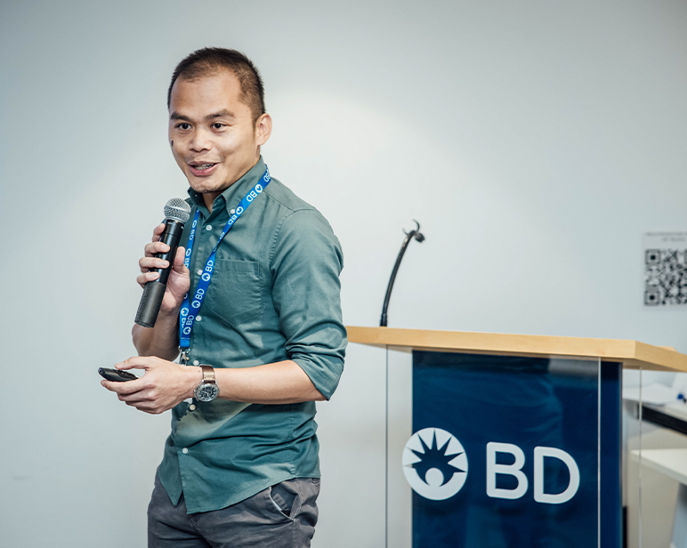 Persona dando una presentación en un podio, sosteniendo un micrófono y un control remoto.