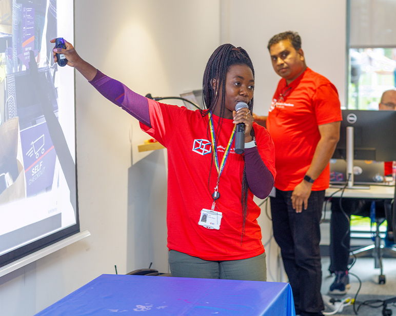 Una persona con una camisa que lleva el logotipo de BD da una presentación utilizando un micrófono y un puntero láser, mientras que otro individuo permanece de pie cerca de un equipo informático.