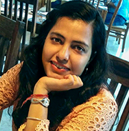 Una persona sentada a una mesa con una mano apoyada bajo la barbilla, vistiendo un top de encaje naranja, en un comedor con sillas de madera.