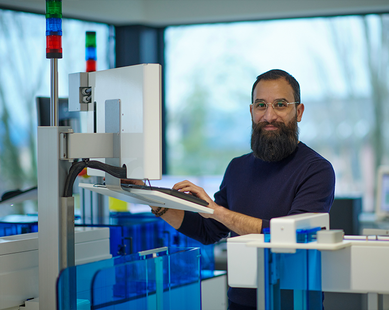 Una persona de pie frente a una estaci&oacute;n de trabajo de automatizaci&oacute;n m&eacute;dica, interactuando con un monitor montado en un entorno de laboratorio con equipos de tonalidad azul.