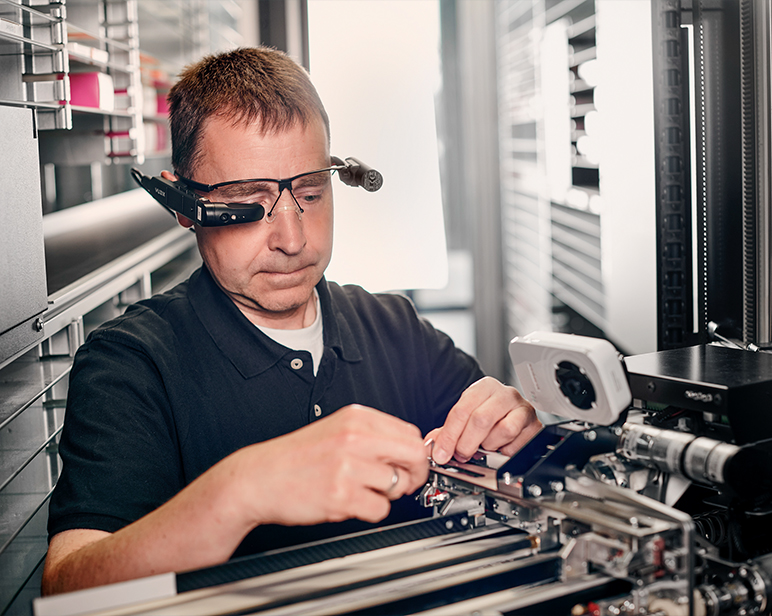 Una persona, vistiendo un polo oscuro, se encuentra sentada en un puesto de trabajo, ajustando componentes en un equipo mec&aacute;nico de precisi&oacute;n.
