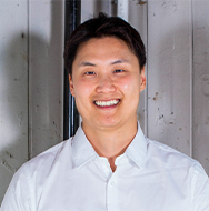 Un individuo con una camisa blanca abotonada, de pie frente a un fondo industrial o metálico claro.