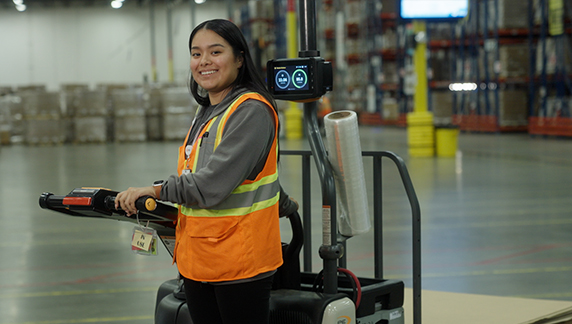 Una persona con un chaleco de seguridad de alta visibilidad, de pie junto a maquinaria de almac&eacute;n motorizada, dentro de un gran centro de distribuci&oacute;n.