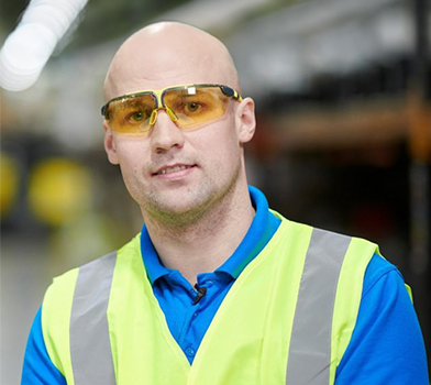 Una persona mirando directamente a la c&aacute;mara, vistiendo un chaleco de seguridad de alta visibilidad y una camisa azul con cuello, en el interior de un almac&eacute;n con estanter&iacute;as de fondo.
