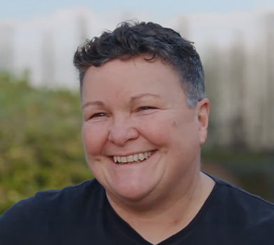 Una persona al aire libre, sonriendo a la c&aacute;mara, vistiendo una camisa oscura, con follaje y cielo borrosos de fondo.