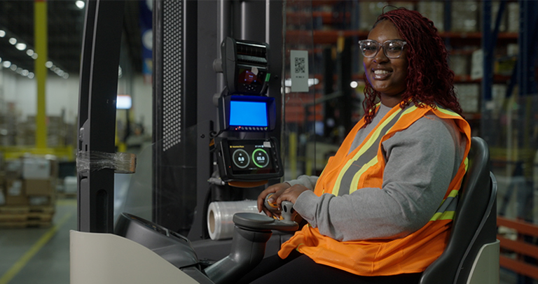 Imagen que muestra a una persona con un chaleco de seguridad de alta visibilidad operando maquinaria motorizada de almacén dentro de un centro de distribución, con pasillos de estanterías y materiales apilados.